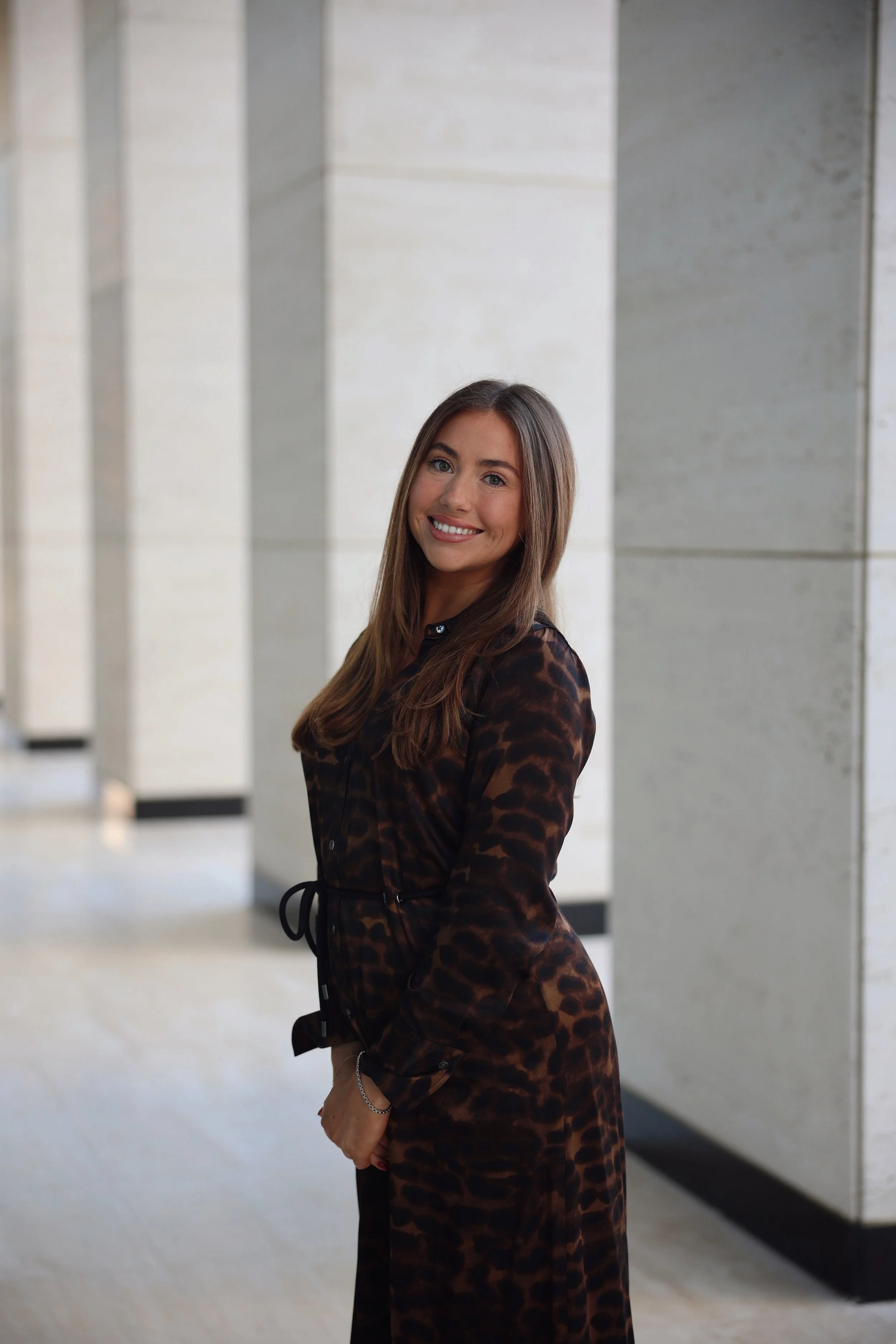 A woman with long brown hair, wearing a dark leopard print dress, smiling and looking at the camera, standing in front of a modern, minimalist concrete wall.