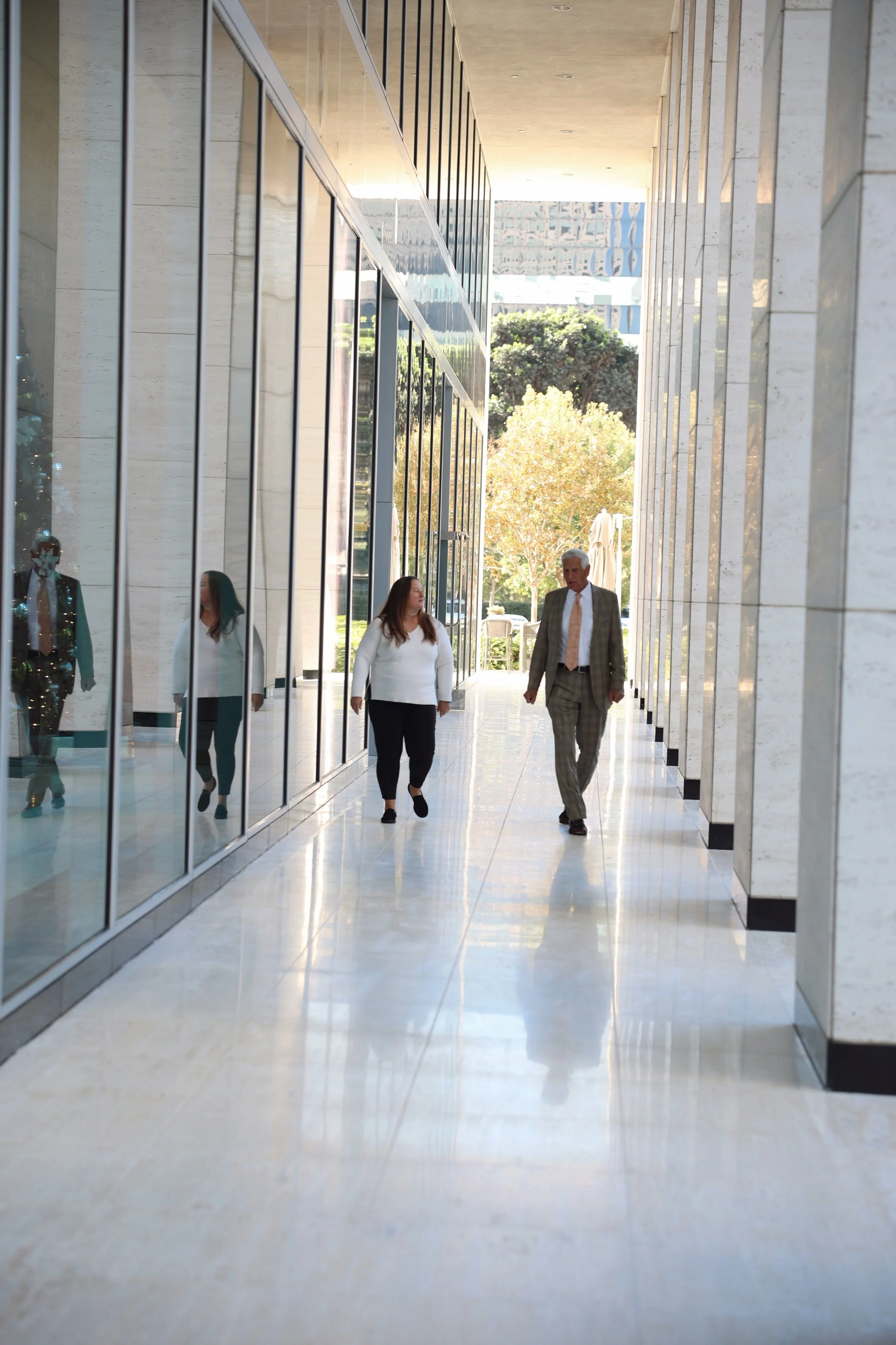 Two people walking through a modern, outdoor corridor with reflective glass walls and trees in the background.