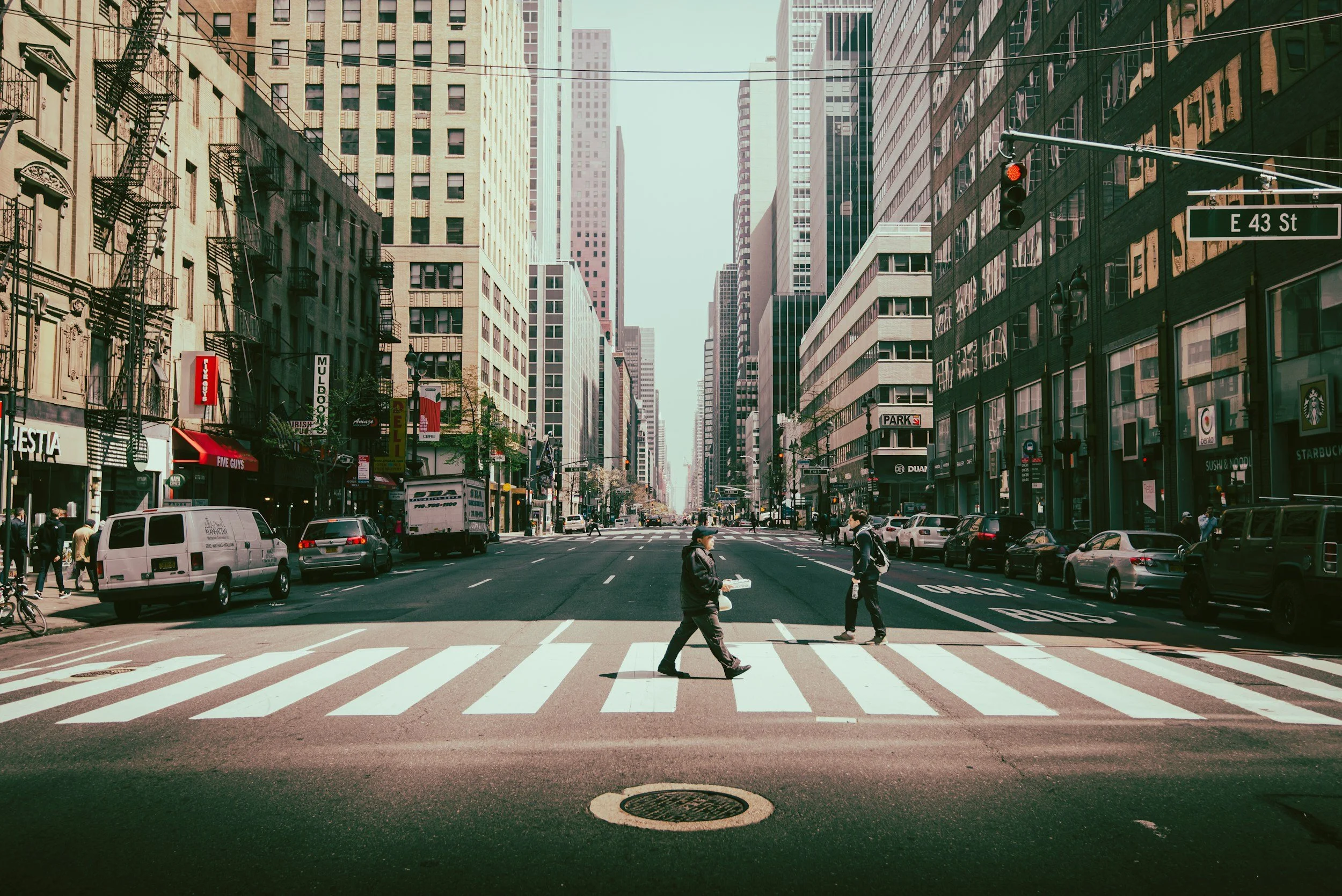 City street scene with pedestrians crossing at a crosswalk, vehicles, and tall buildings on both sides, including a sign indicating E 43 Street.