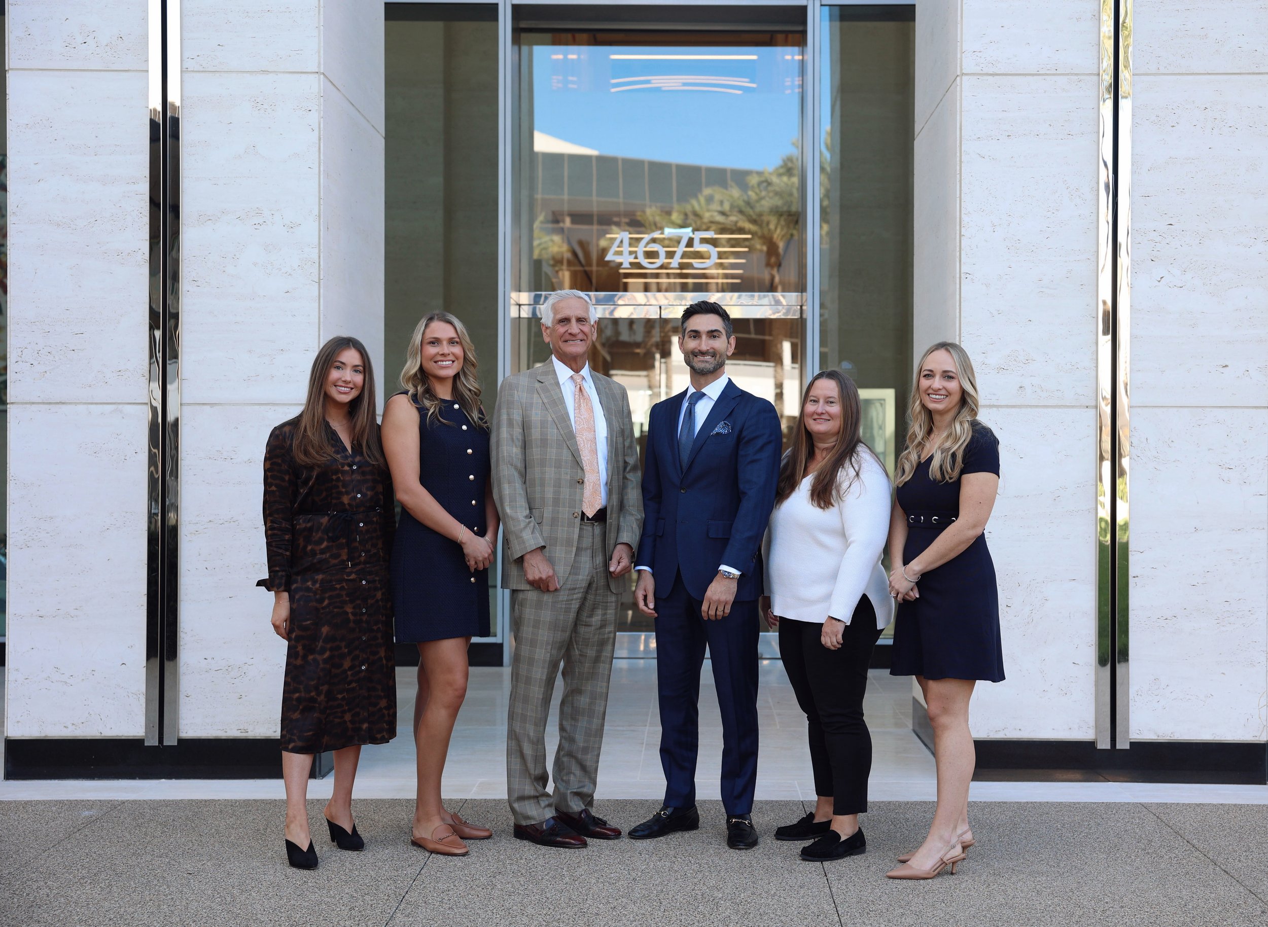 Group of six professionally dressed people standing in front of a building with the address 4675 displayed on the glass door.