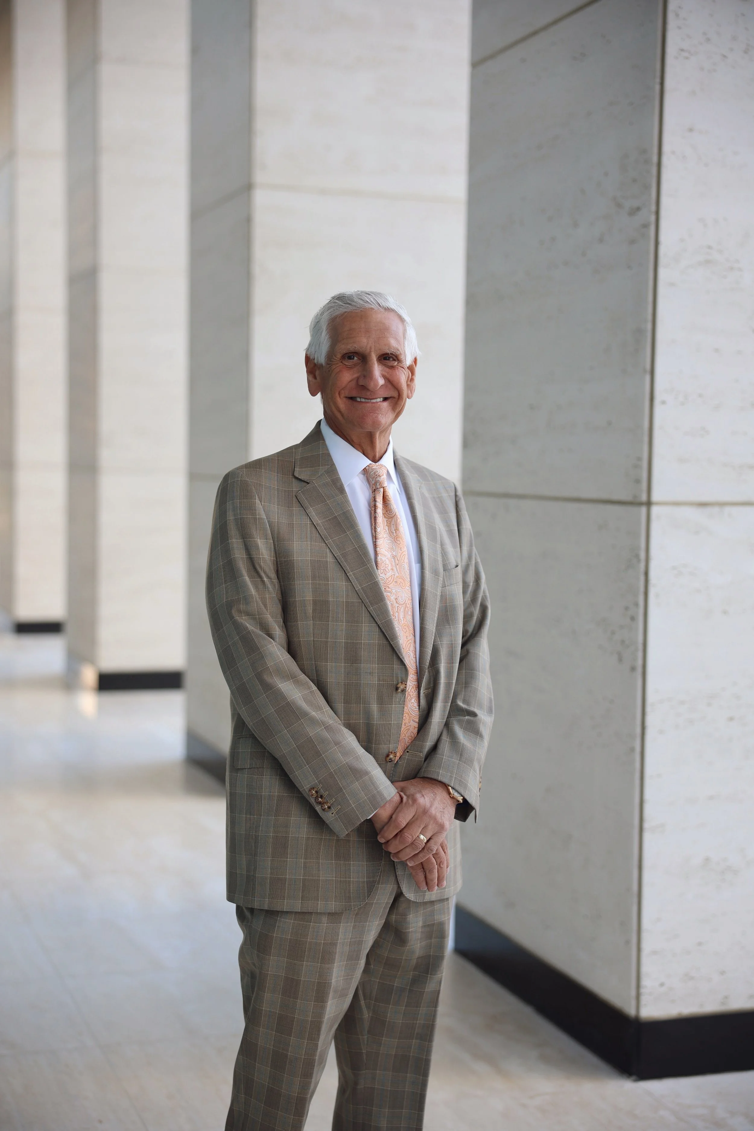 A smiling older man in a light brown plaid suit, white shirt, and pastel tie, standing indoors near a modern light-colored stone wall.