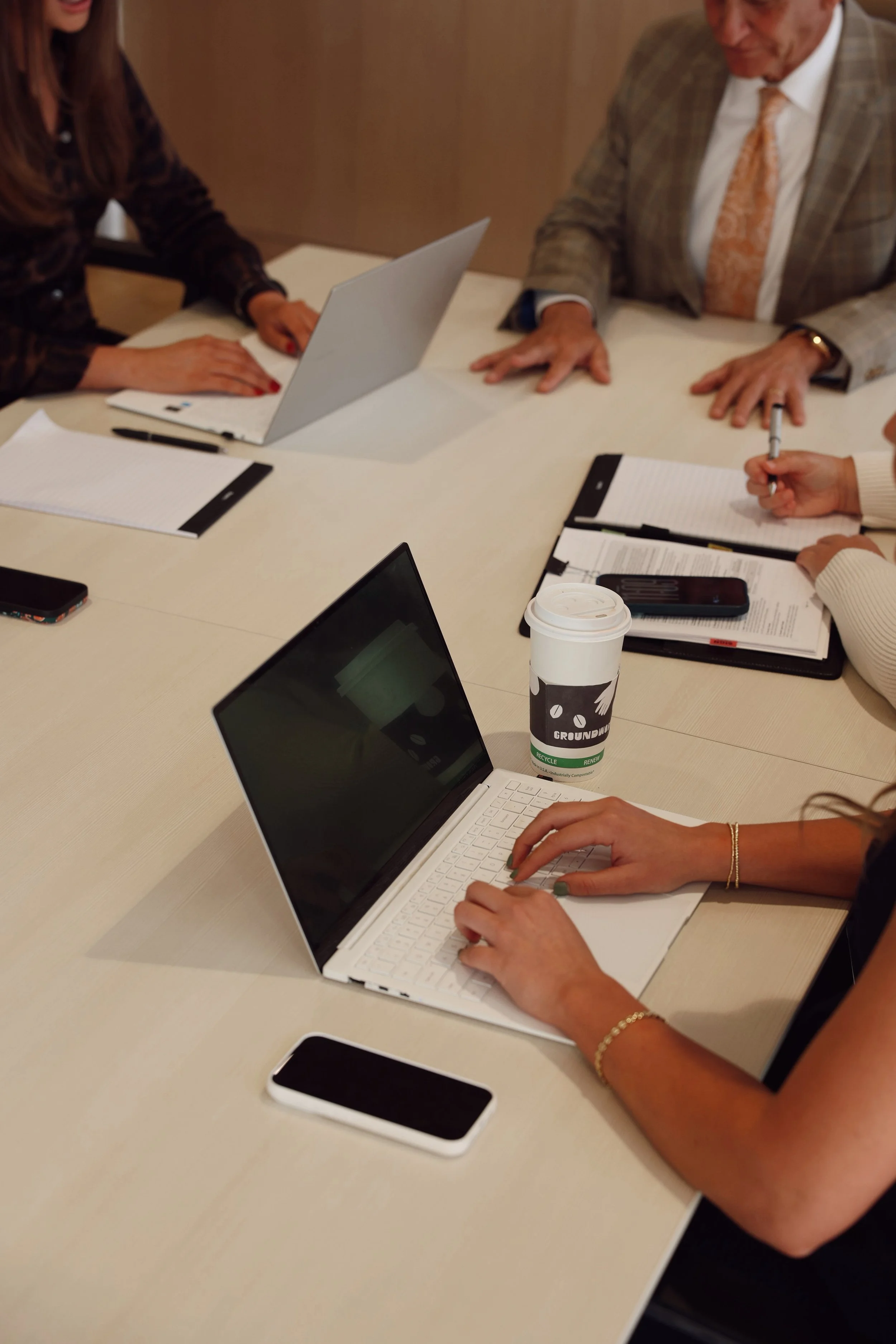 People sitting at a table working on laptops and taking notes, with a disposable coffee cup and smartphones on the table.