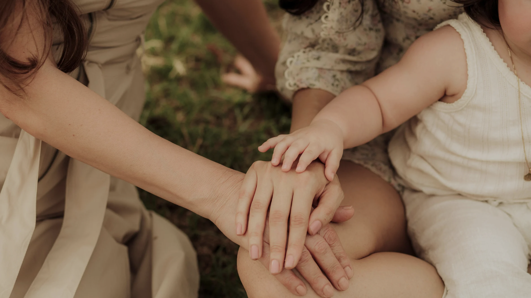 Multiple hands of adults and children stacked together, outdoors on grass, symbolizing unity and togetherness.