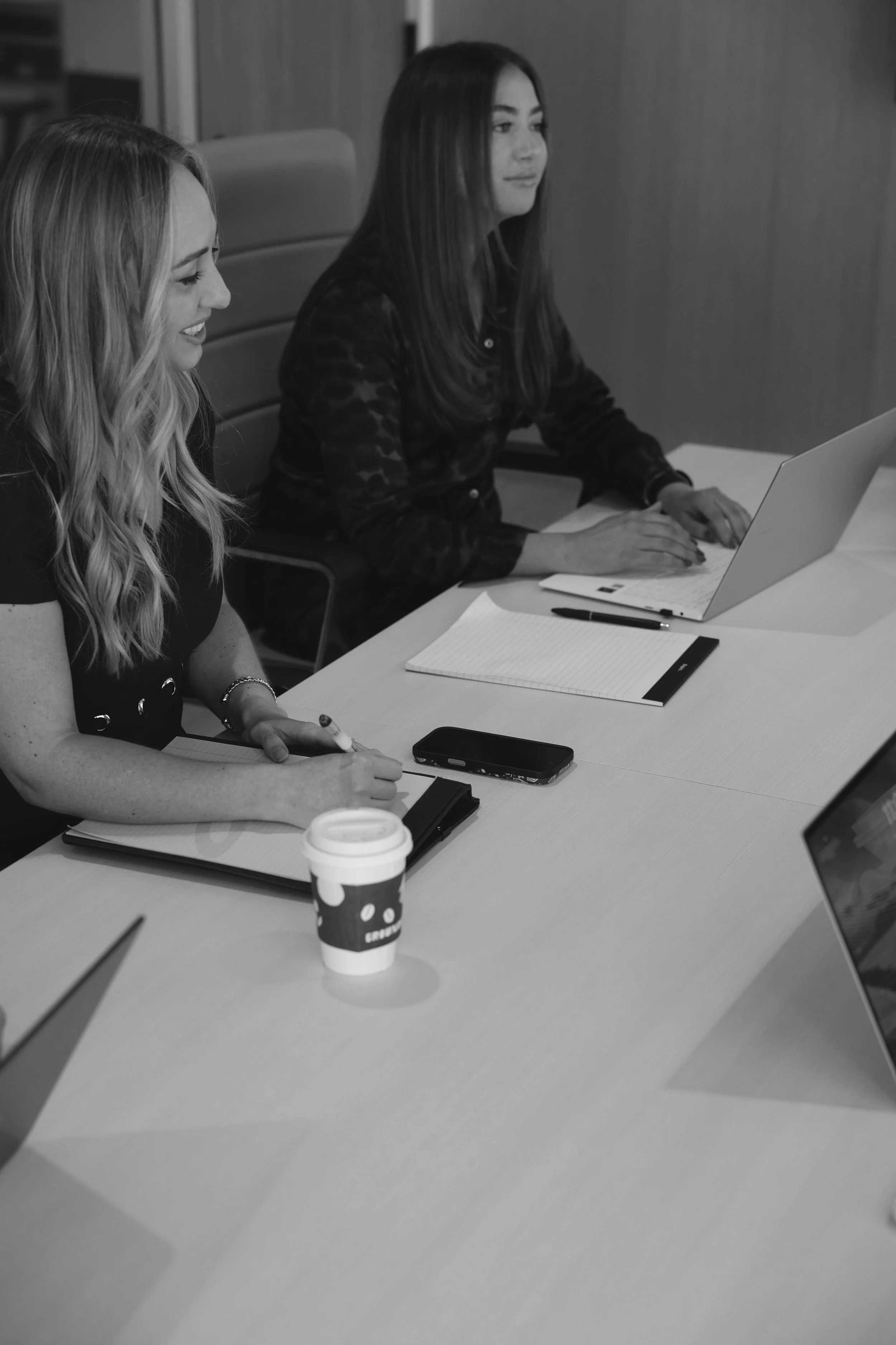 Two women sitting at a conference table in a meeting room, working on laptops, with notebooks, pens, and a coffee cup on the table.