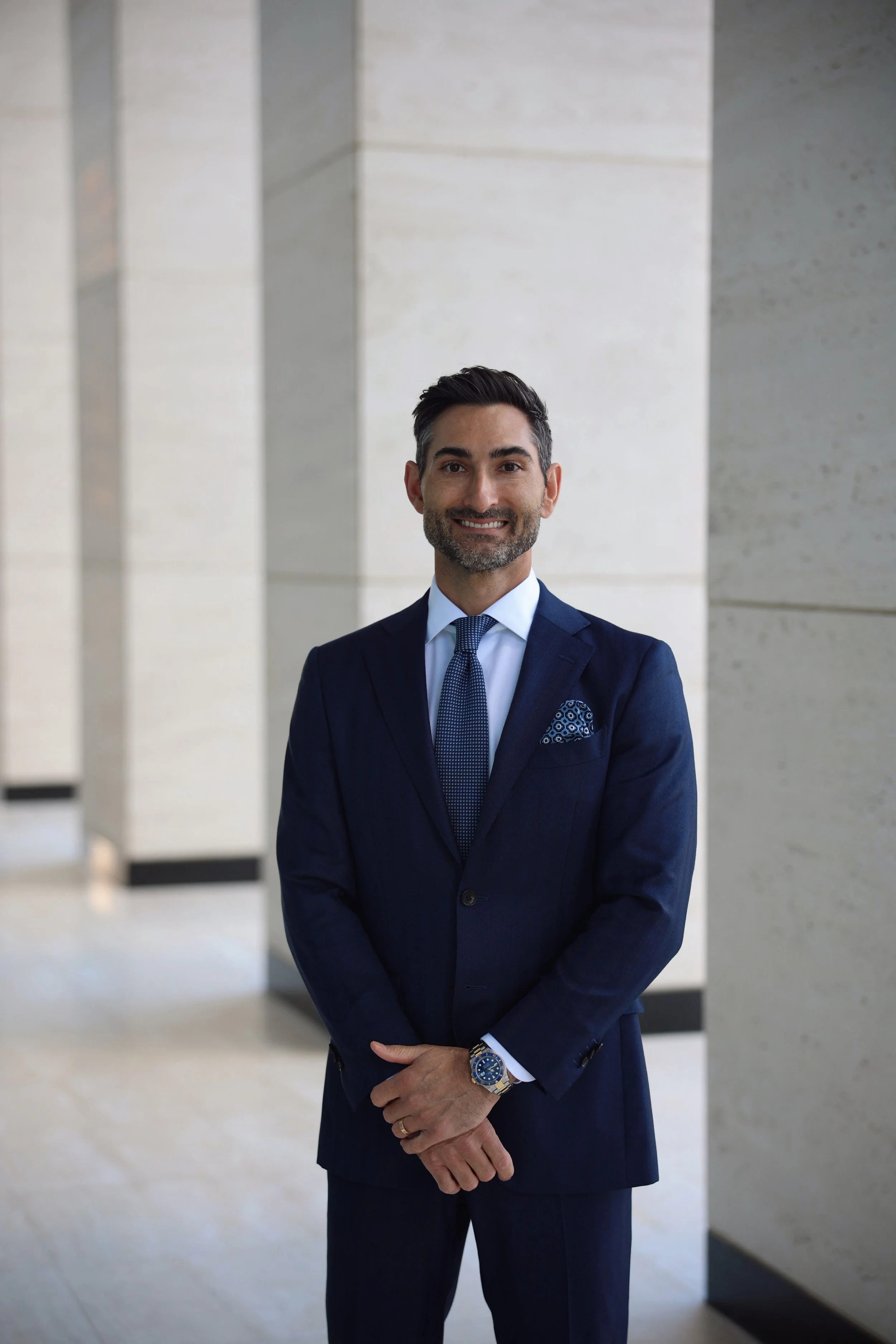 A man in a navy suit standing with arms crossed, smiling, in a modern, neutral-colored indoor space with marble walls.