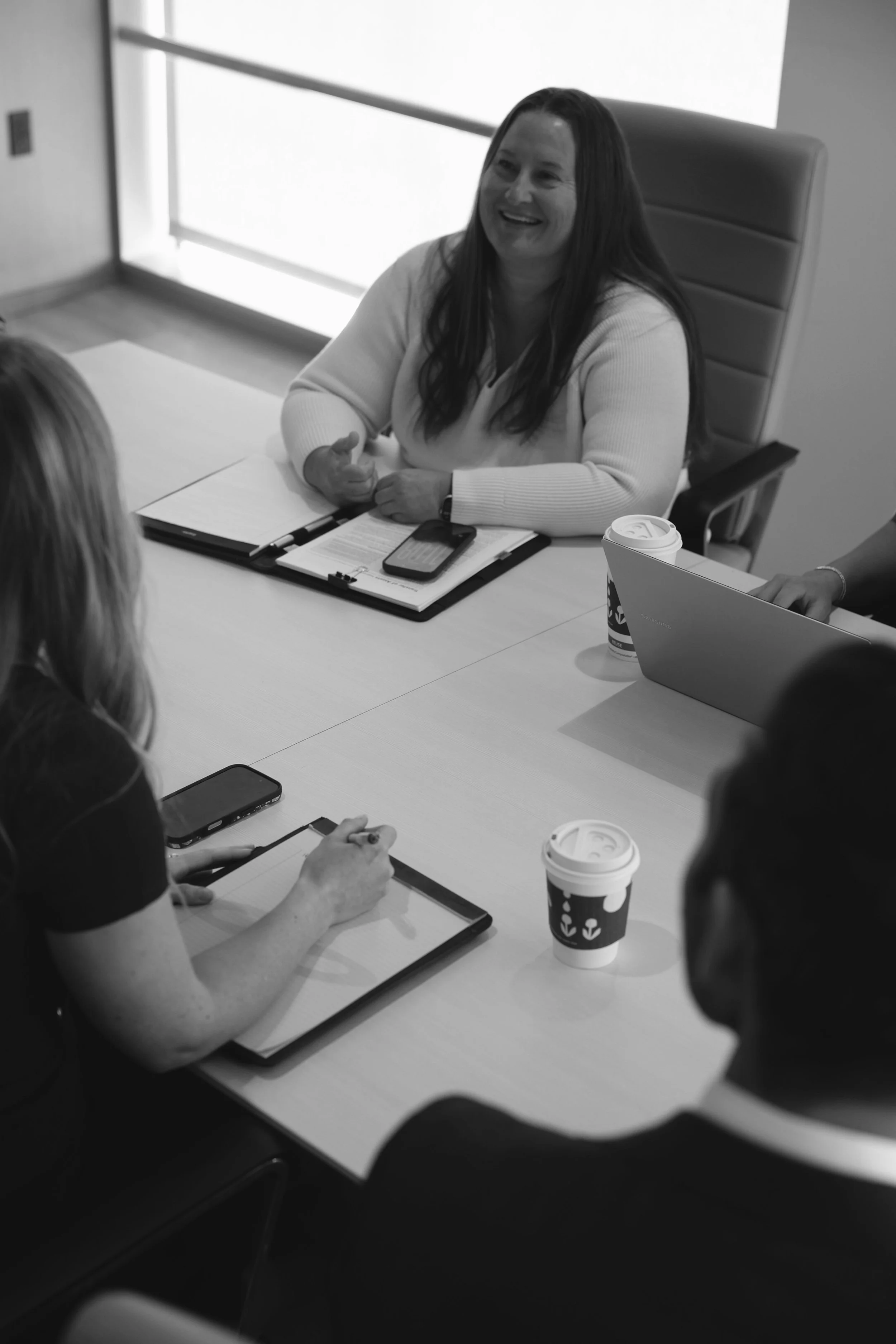 A woman seated at a conference table, smiling and gesturing with her hand during a meeting. There are paper notebooks, a smartphone, and disposable coffee cups on the table, with a window providing natural light.