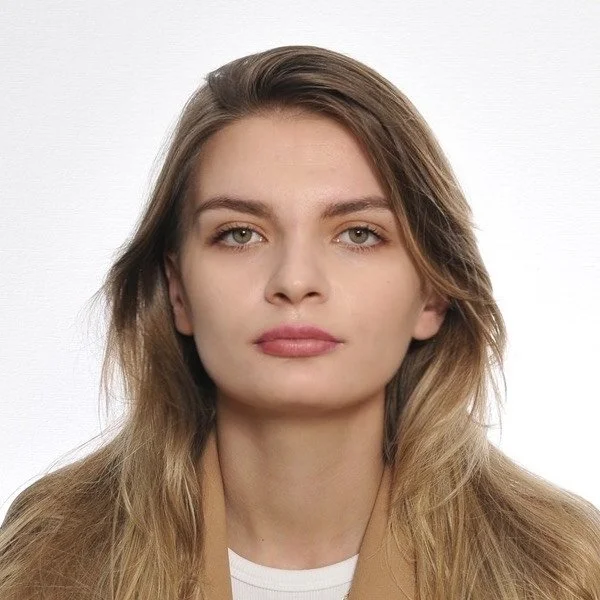 Close-up portrait of a young woman with long, wavy blonde hair, wearing a tan blazer and white top, against a plain white background.