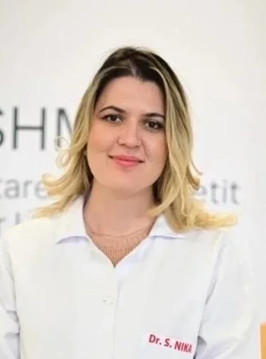 A woman with shoulder-length blonde hair, wearing a white lab coat with the name tag 'Dr. S. NIKA', standing in front of a light-colored background with partial text.