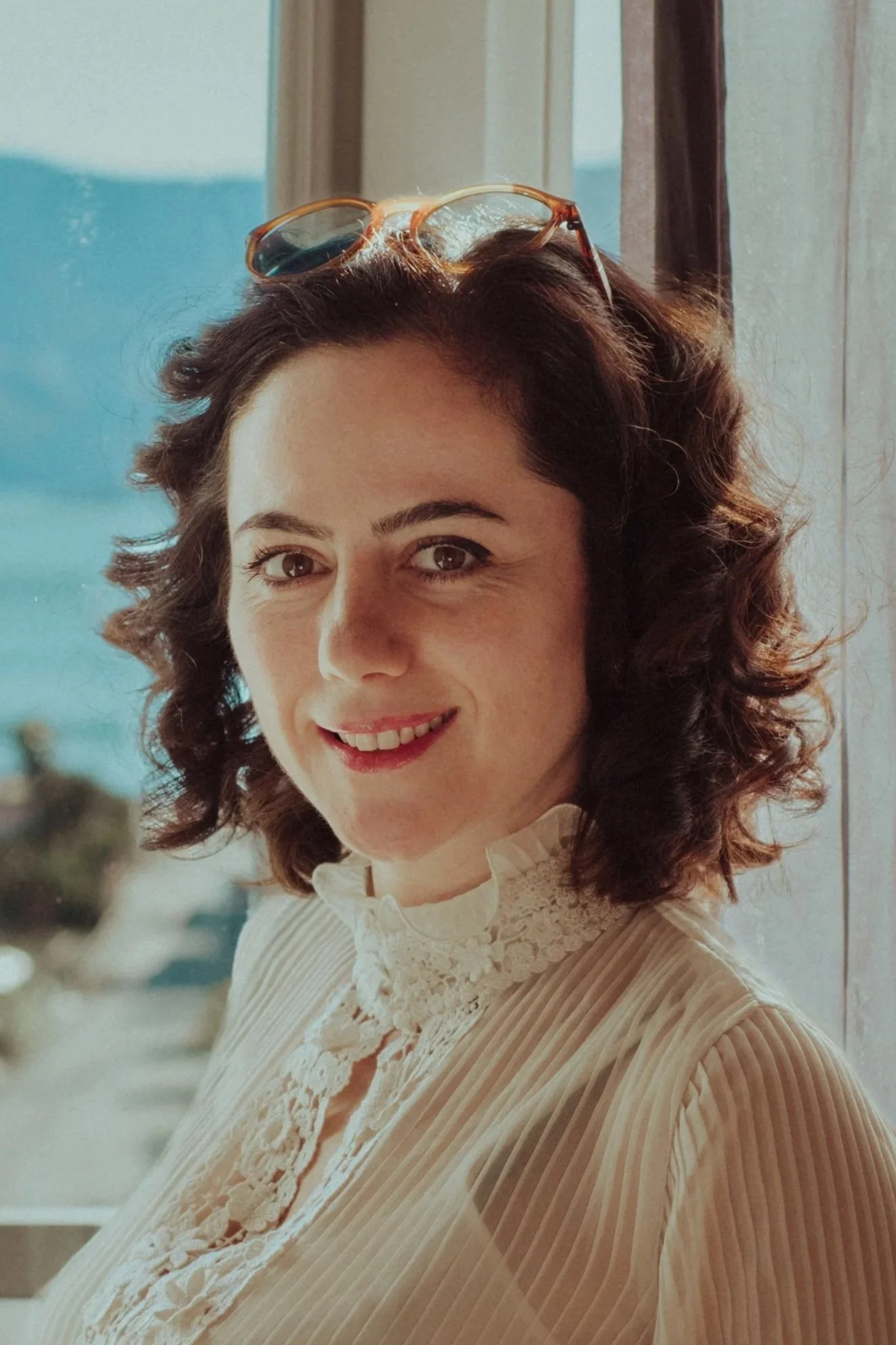 A woman with dark curly hair wearing glasses on her head, smiling while sitting near a window with a view of the sky and outdoors, dressed in a light-colored blouse with lace details.