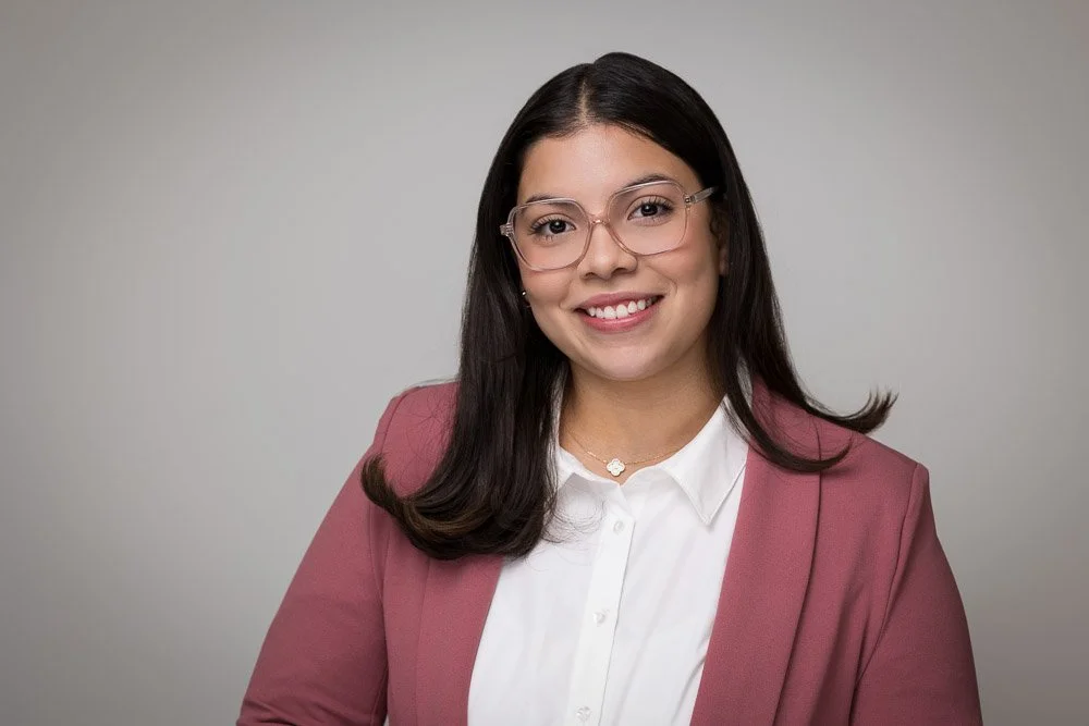 Professional woman with dark hair, glasses, white blouse, and pink blazer smiling at the camera against a plain gray background.