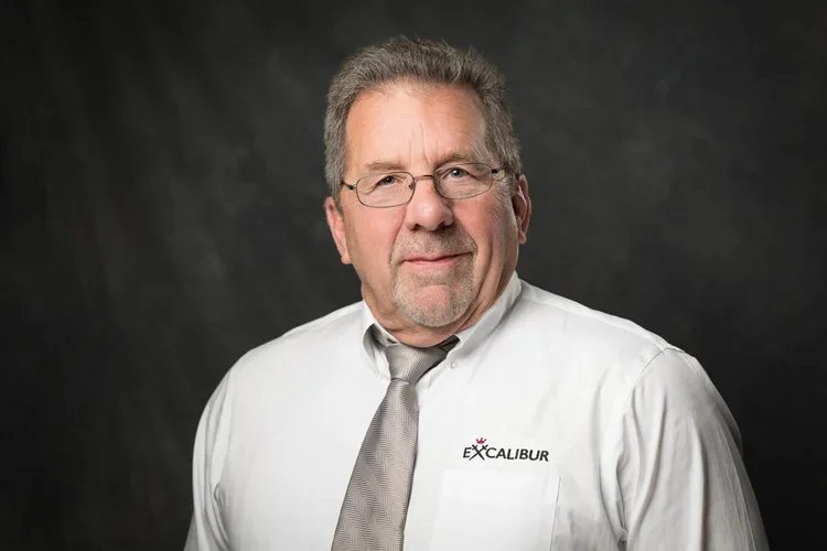 A middle-aged man with gray hair and glasses wearing a white dress shirt and a gray tie, standing in front of a dark background.