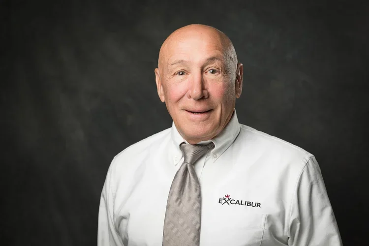 A bald man wearing a white dress shirt with a Caliburn logo and a gray tie, smiling against a dark gray background.