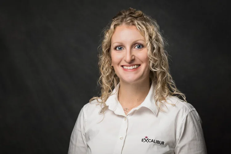 A woman with shoulder-length curly blonde hair smiling, wearing a white button-up shirt with a logo and the word 'EXCALIBUR' on it, against a dark background.