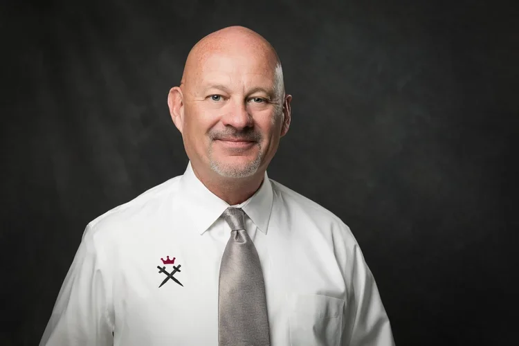 A man with a bald head and beard wearing a white shirt, gray tie, and a red and black logo on his shirt, standing against a dark gray background.