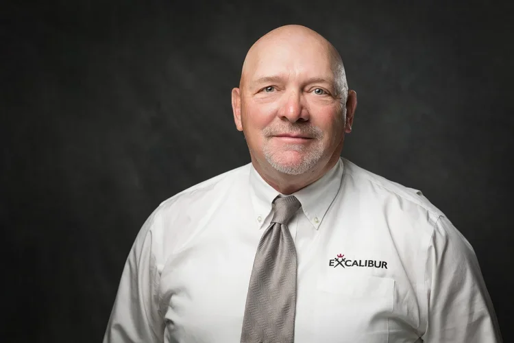 A middle-aged man with a bald head and a gray goatee, wearing a white shirt with a logo that says 'EX CALIBUR' and a gray tie, standing against a dark background.