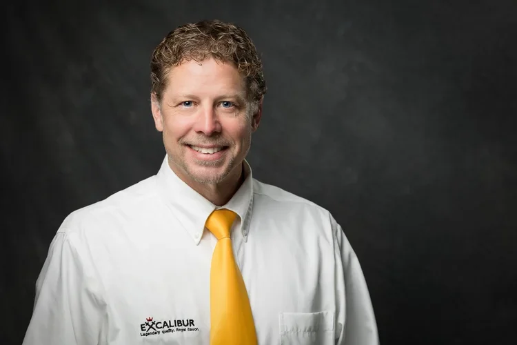 Man with curly hair smiling, wearing a white shirt with a yellow tie, and a logo that says Excalibur.