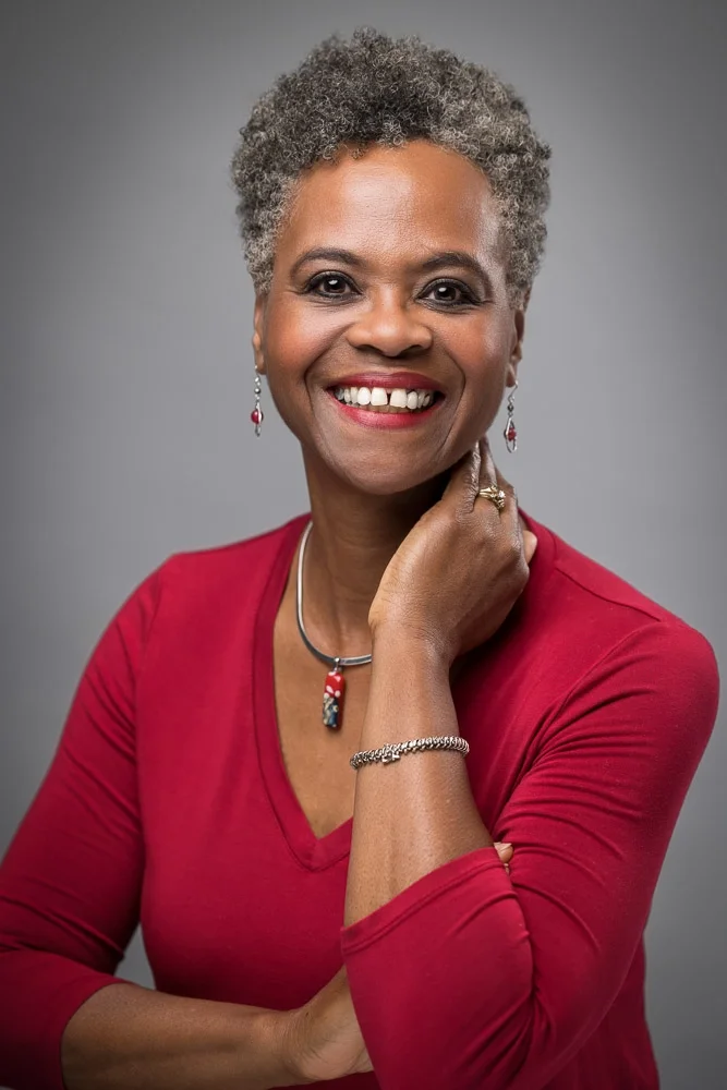 A smiling middle-aged African American woman with short gray curly hair wearing a red top and jewelry, posing against a plain gray background.