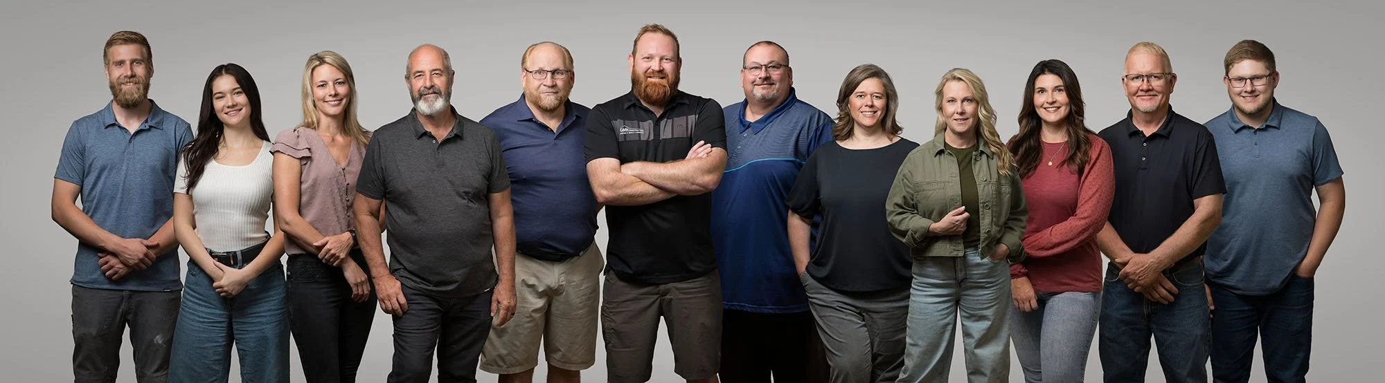 Group of fourteen diverse adults standing together against a light gray background, smiling for a team photo.