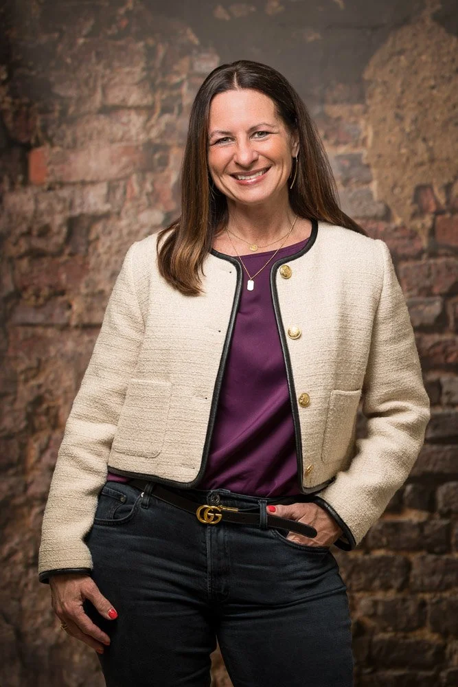 A woman with long brown hair, wearing a beige blazer with gold buttons, a purple blouse, black pants with a Gucci belt, standing in front of a brick wall, smiling at the camera.