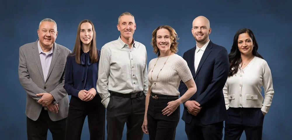 Six diverse professionals standing in a row in front of a blue background, dressed in business attire, smiling at the camera.