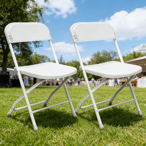 Two white folding chairs on green grass outdoors with trees and a partly cloudy sky in the background.