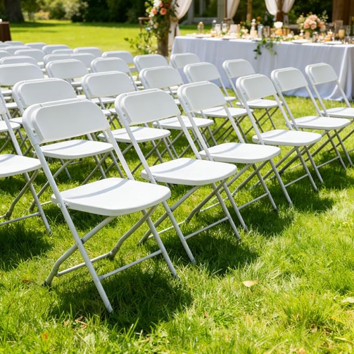 Empty white folding chairs arranged outdoors on green grass at a wedding or event ceremony with decorated table in the background.
