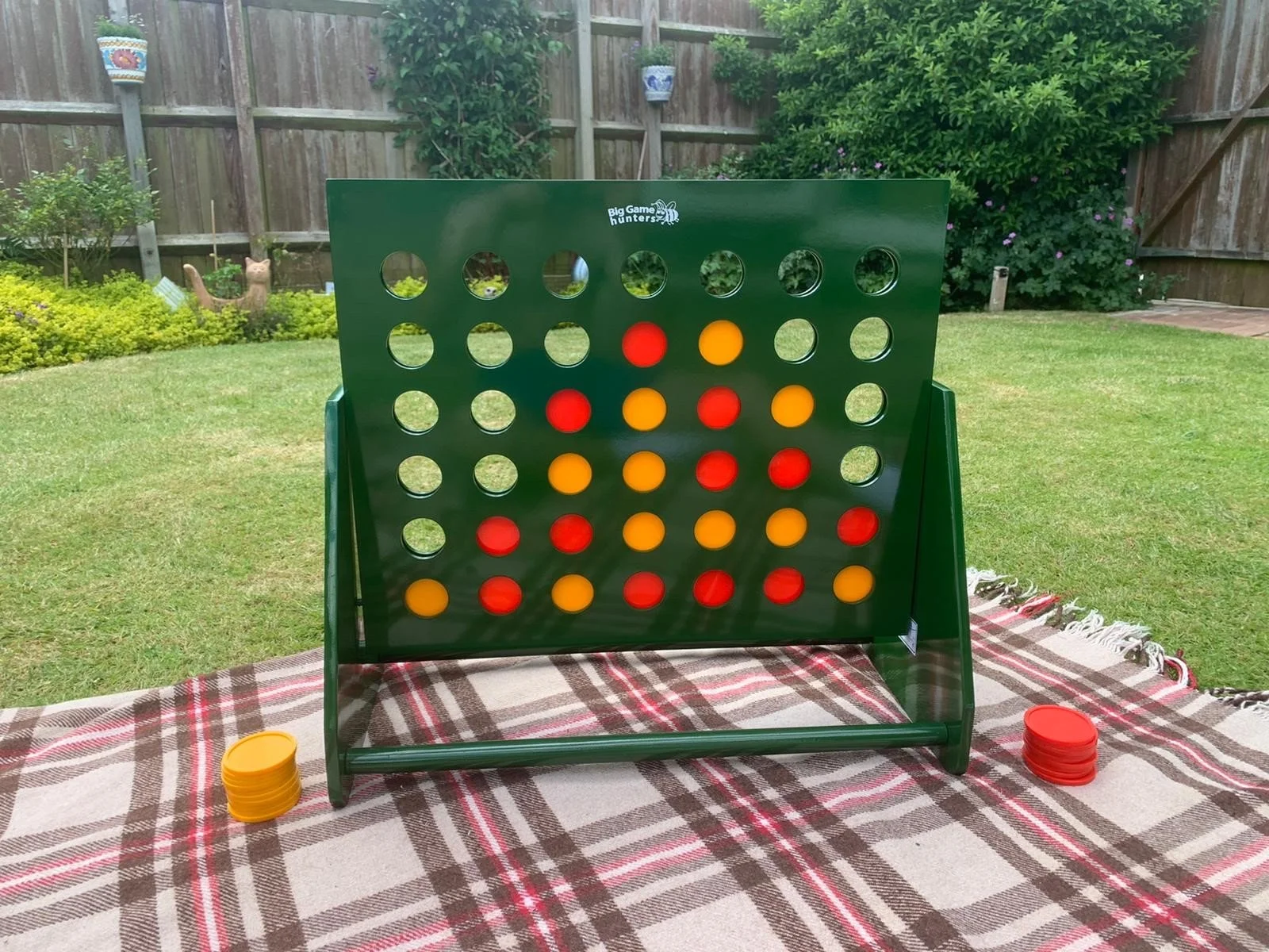 A game of Connect Four set up outdoors on a picnic table with a patterned tablecloth. The game board is green with red and yellow discs, some in the slots. Two stacks of discs, one red and one yellow, are on either side of the game board. The background shows a backyard with grass, bushes, a fence, and garden decorations.
