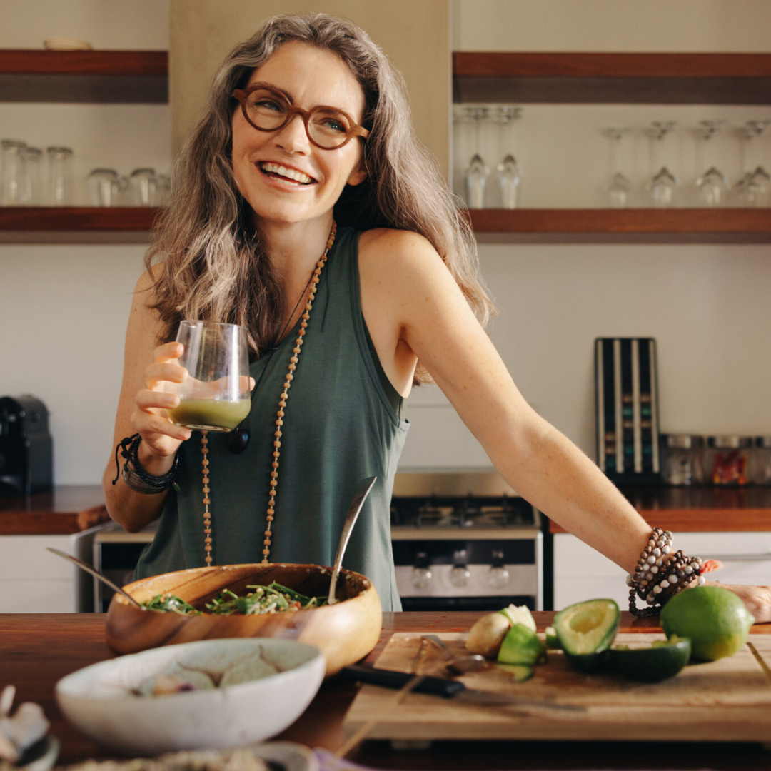 Evidence-Based & Integrative:Combining the precision of modern medicine with the art of holistic healing. Img of a woman with gray hair & glasses holding a smoothie, wearing a sleeveless green top & beaded jewelry, a bowl of salad on the counter.