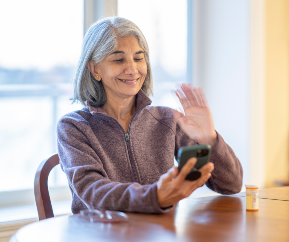 Smile woman with gray hair waving while looking at her phone with a medicine bottle on the table.