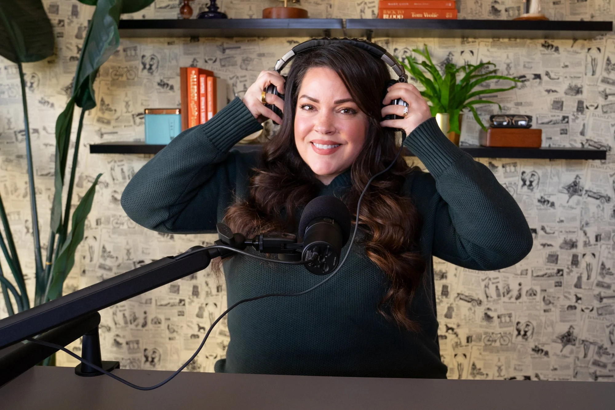 A woman with long, dark, curly hair smiling while wearing headphones in front of a microphone, in a room with a patterned wallpaper background and shelves with books and decor.