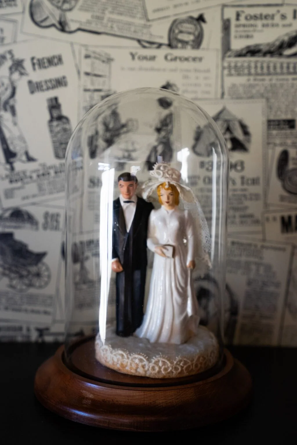 A wedding figurine under a glass dome, featuring a bride in a white dress with a veil and a groom in a black tuxedo, placed on a wooden base with a vintage newspaper background.