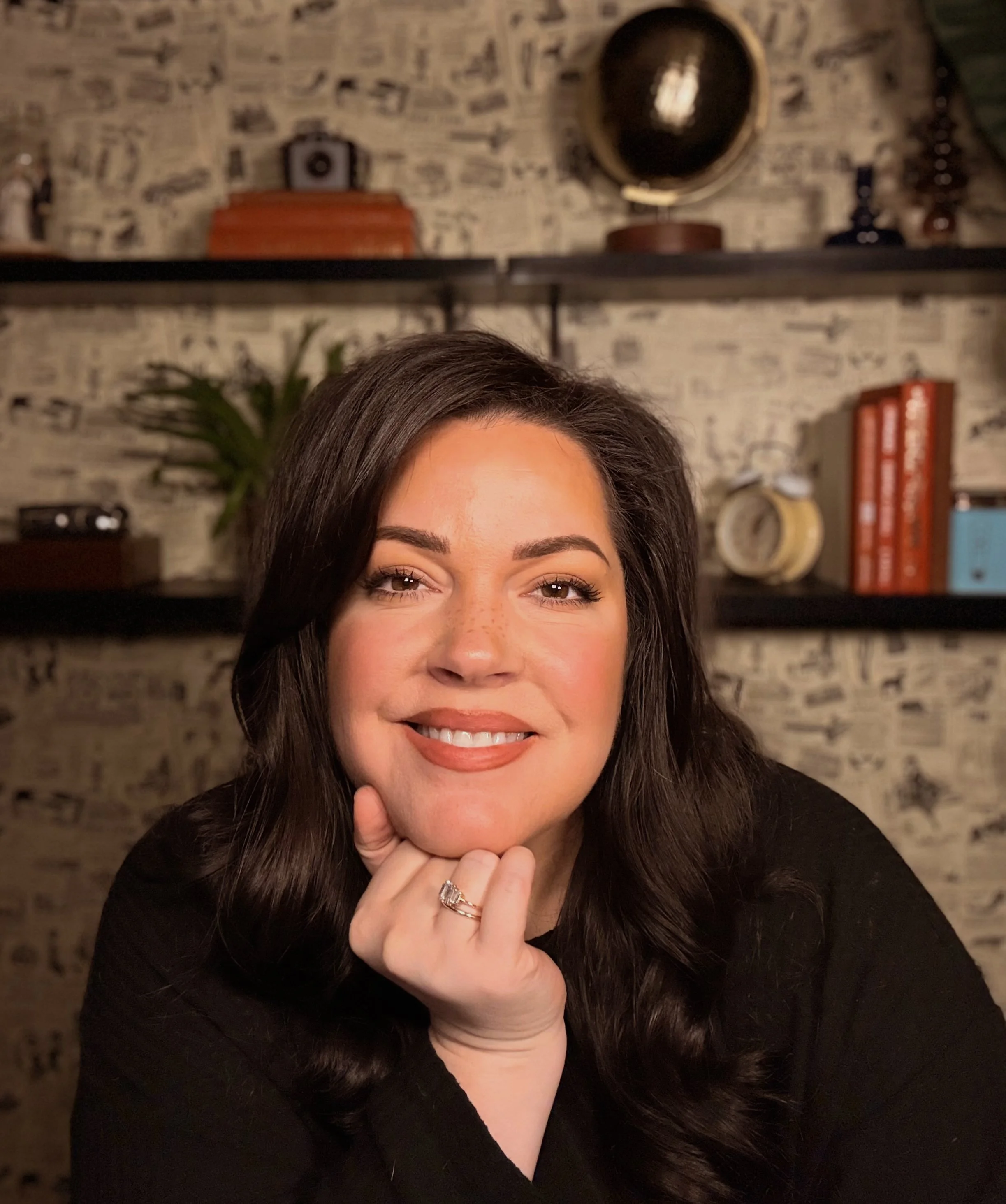 Close-up of a smiling woman with long dark hair resting her chin on her hand, wearing a black top, in front of a background with shelves, books, and decorative items.