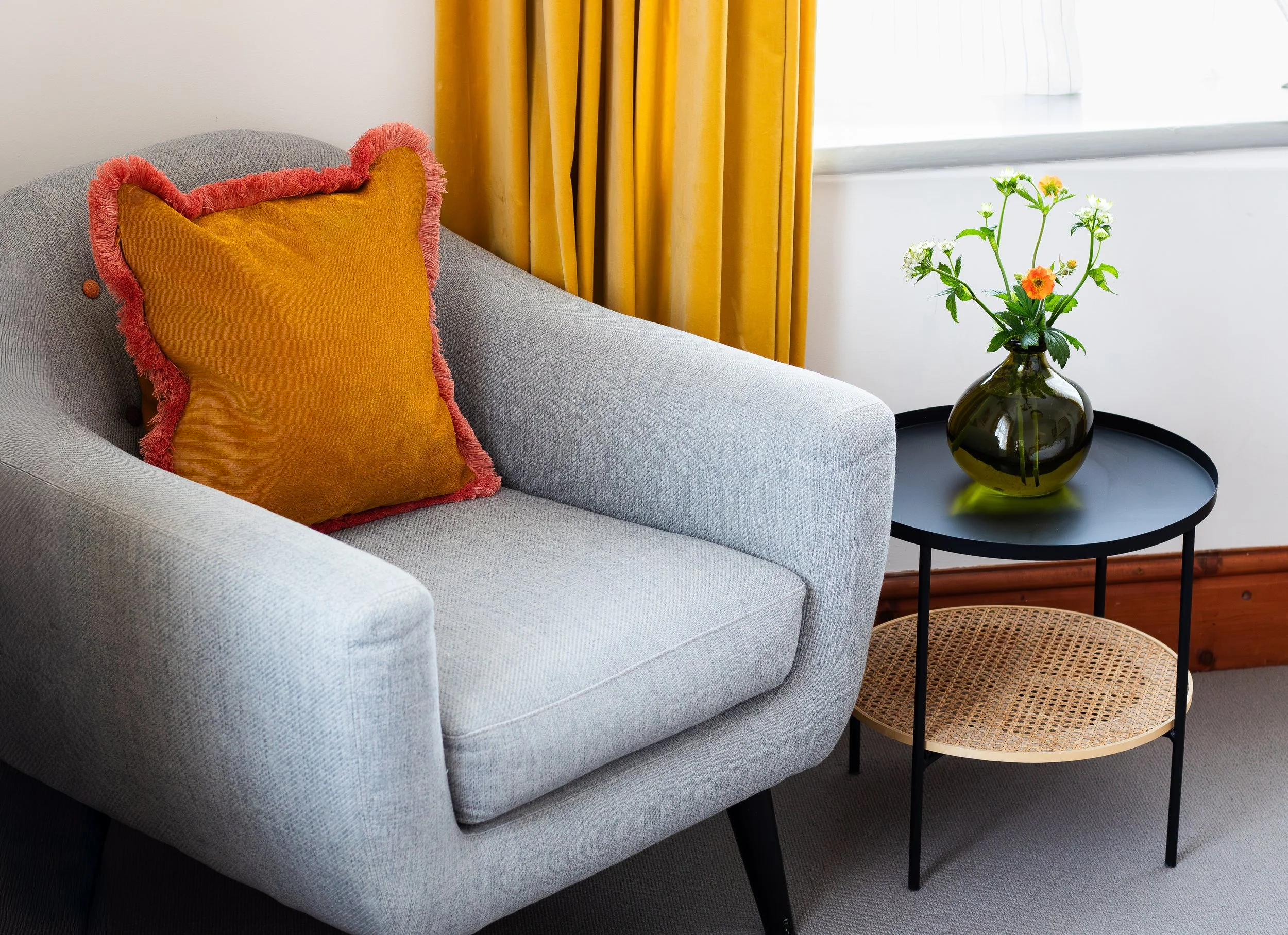 A light gray upholstered sofa with a large mustard yellow pillow with a pink fringed border, beside a black round side table holding a dark green vase with white and orange flowers. Yellow curtains and a window are in the background.