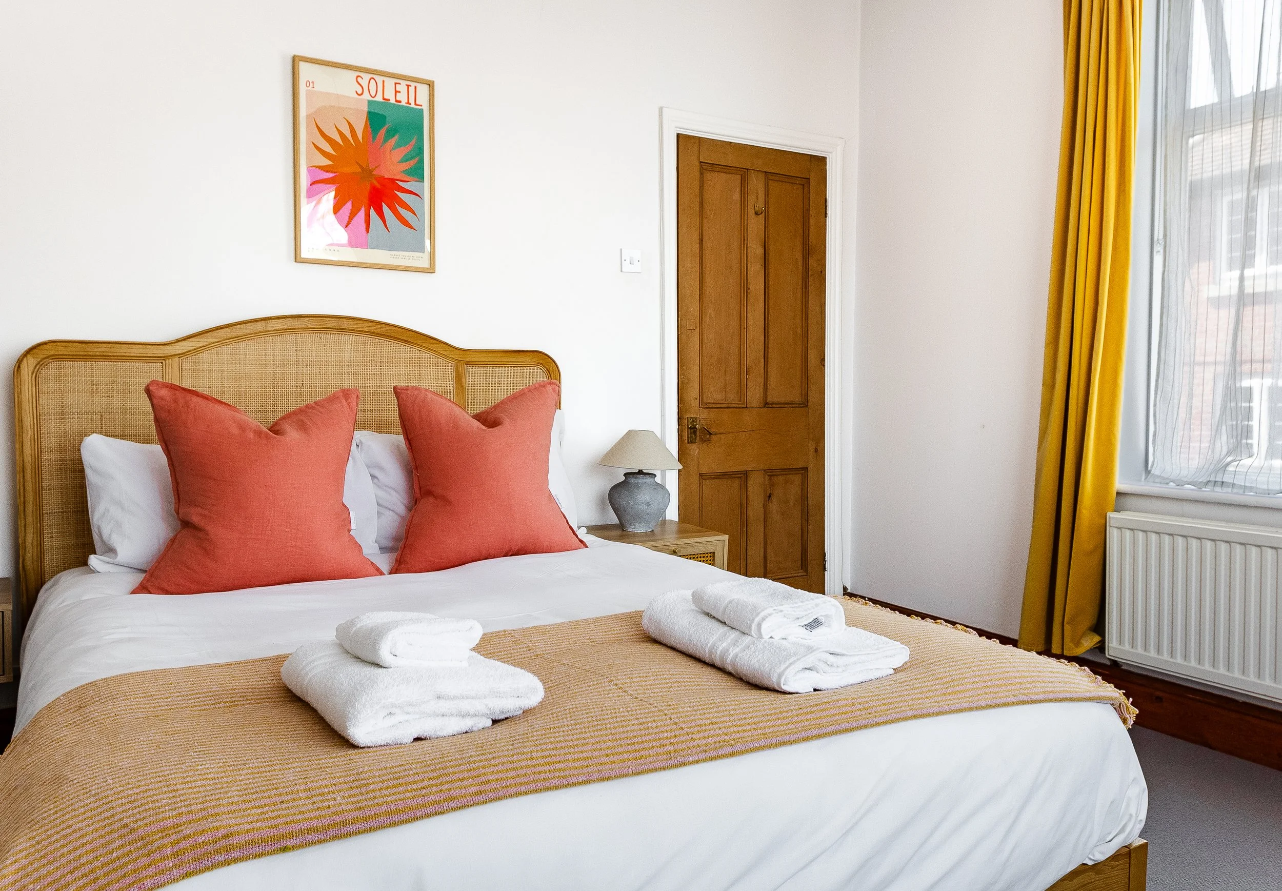 A neatly made bed with white sheets, an orange-striped bed runner, and three large coral-colored pillows on a wooden bed frame. Towels are placed at the foot of the bed. There is a small bedside table with a gray lamp. A colorful framed art print tit