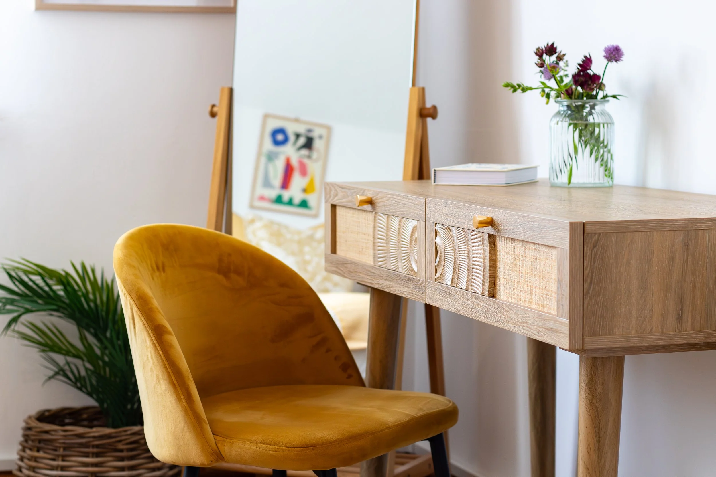 A wooden desk with a small floral arrangement, a book, and a mirror, partially visible yellow chair, and a green potted plant in a woven basket nearby.