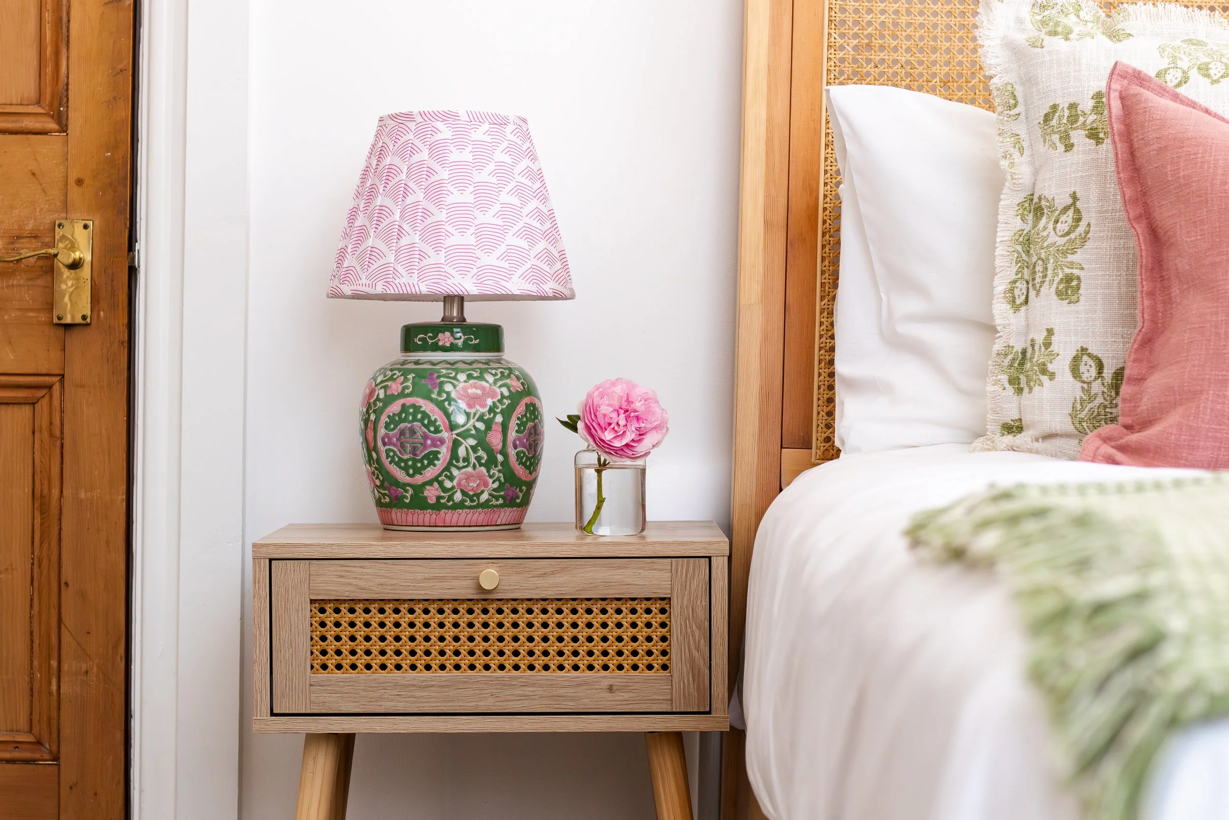 A close-up of a beige wooden nightstand with rattan detail on the drawer front, beside a bed with white and pink pillows, a green ceramic lamp with pink and white floral patterns and a pink lampshade, and a small glass vase with pink peony flowers on