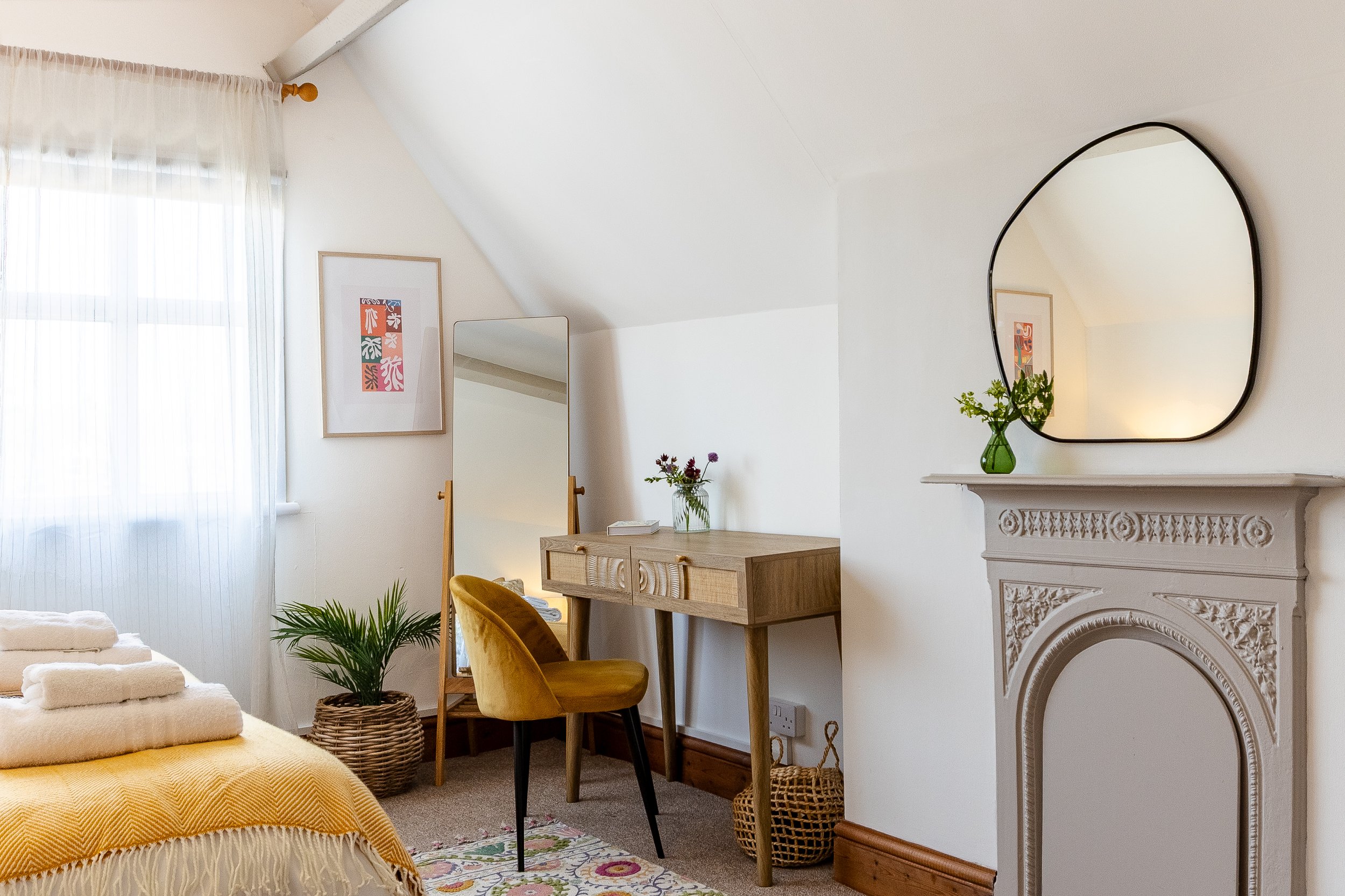 A cozy bedroom corner featuring a white fireplace with intricate detailing, a green vase with flowers, a wooden vanity with a mirror, a yellow velvet chair, and a framed colorful abstract art piece on a white wall. Light filters through sheer curtain