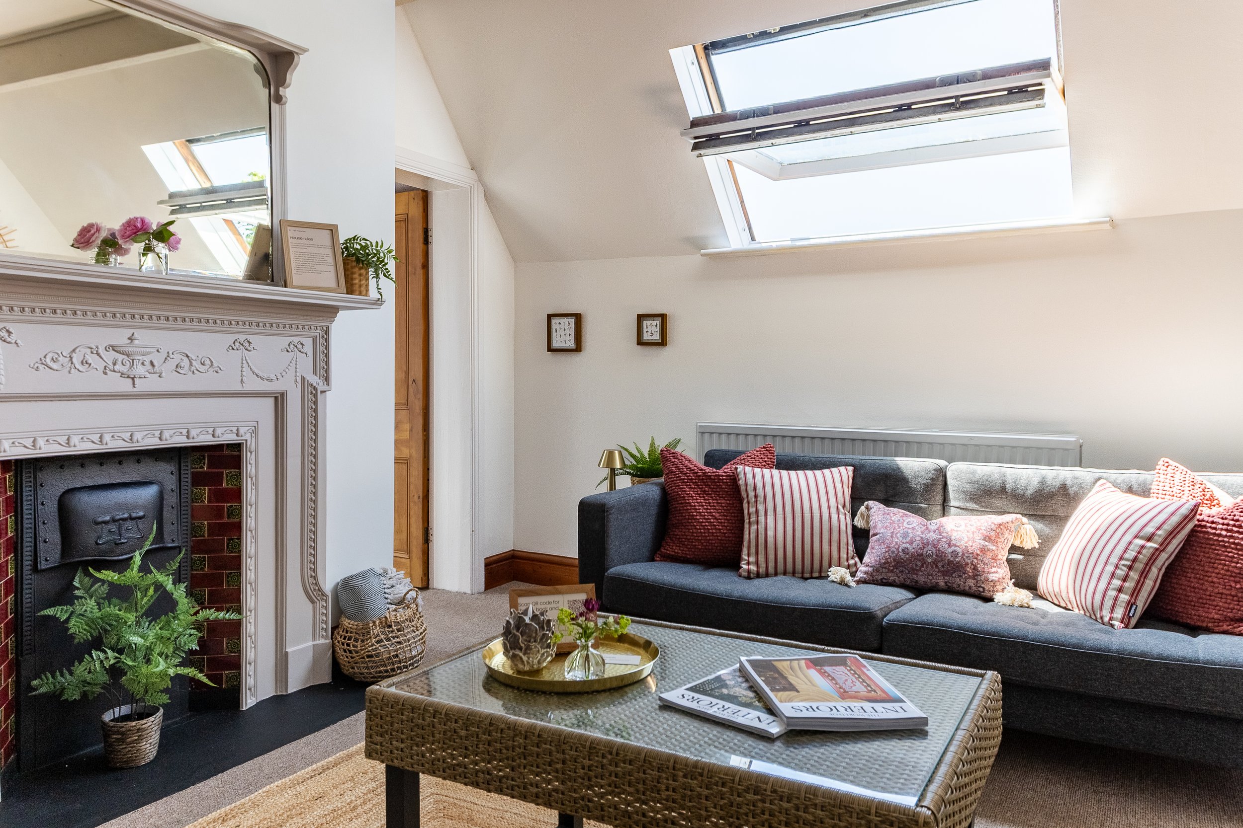 Living room with a large dark gray L-shaped sofa with striped and textured throw pillows, a bamboo coffee table with magazines, a white fireplace mantel decorated with pink flowers and a framed document, a small side table with a brass lamp, and a wo