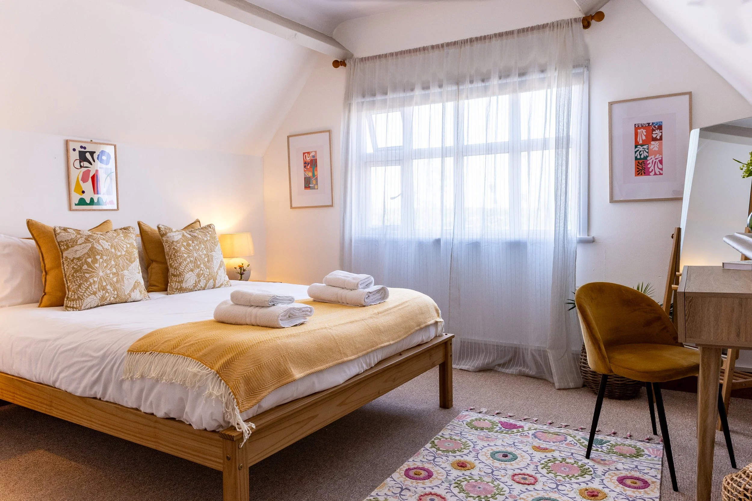 A cozy bedroom with a wooden bed frame, white bedding, and decorative pillows. There are three folded towels on the bed. The room has a large window with sheer curtains, framed artwork, a small lamp, a patterned rug, and a wooden chair.