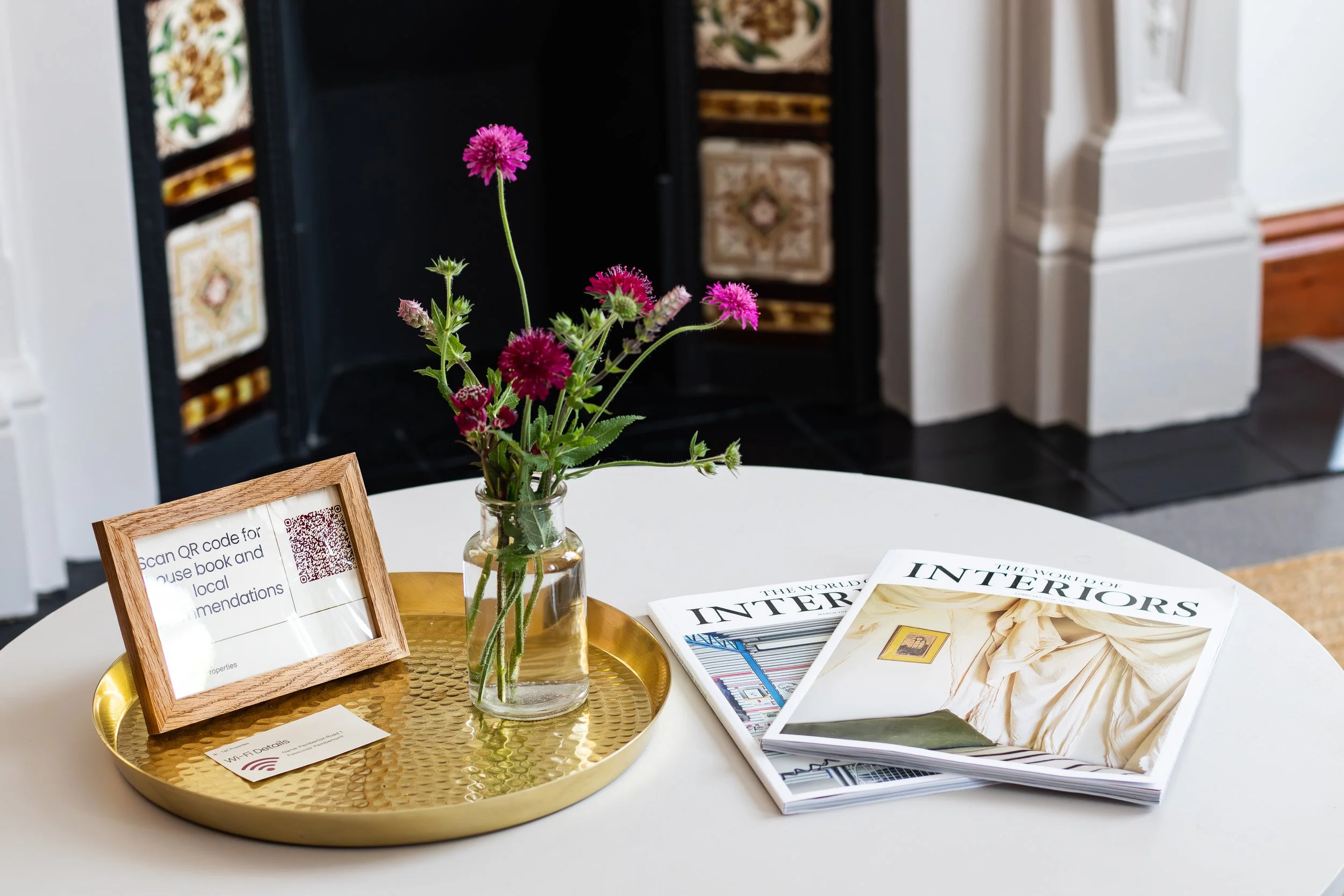 A white round table with a gold tray holding a glass jar of pink and purple flowers, a framed QR code sign, and a booklet titled 'The World of Interiors.' In the background, there's a black fireplace with decorative tiles and part of a wooden floor.