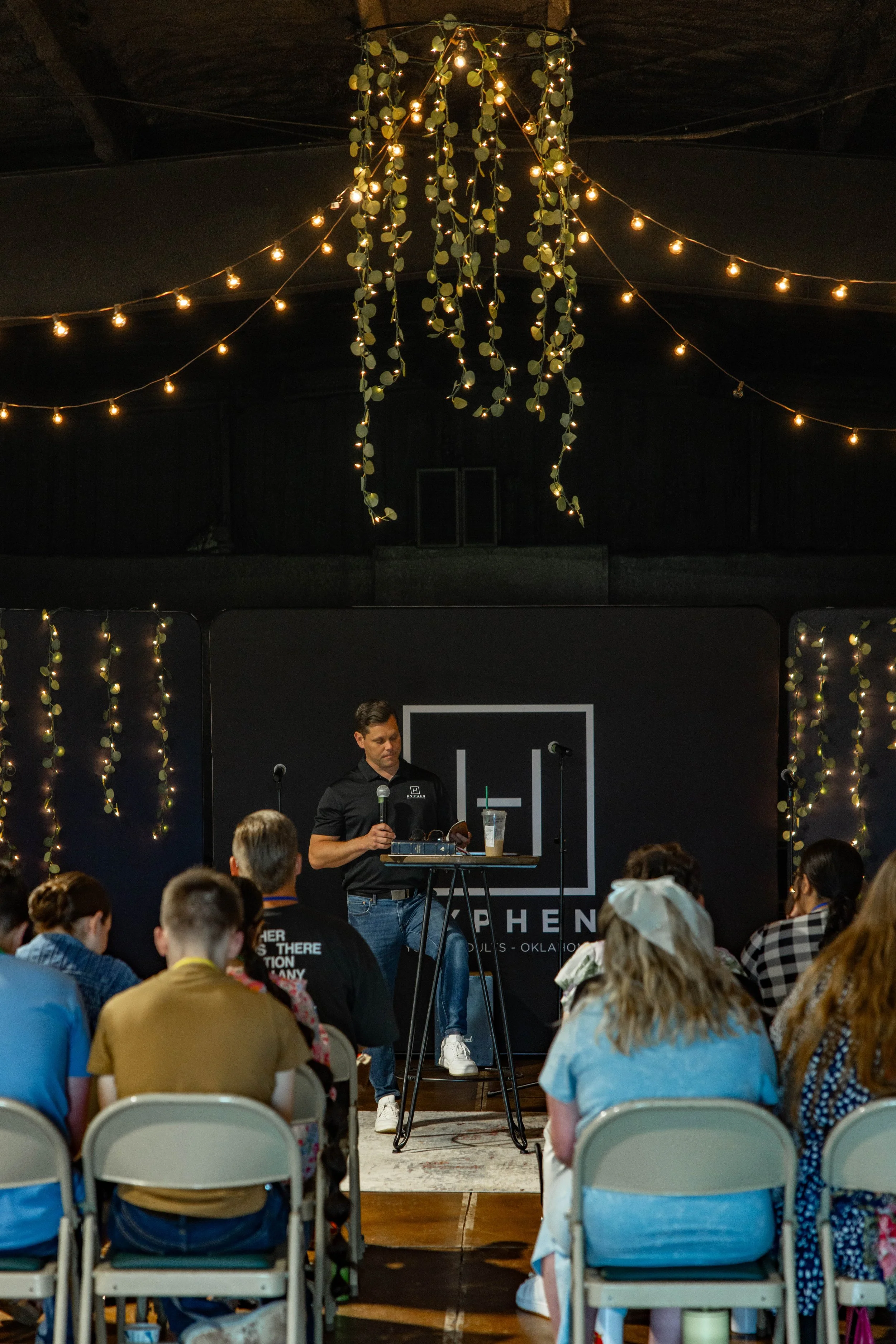 A man in black shirt standing at a podium with a microphone in front of an audience, in a room decorated with hanging string lights and greenery.