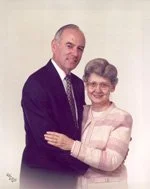 A man and an elderly woman standing close together, smiling, with the man in a dark suit and the woman in a pink striped sweater.