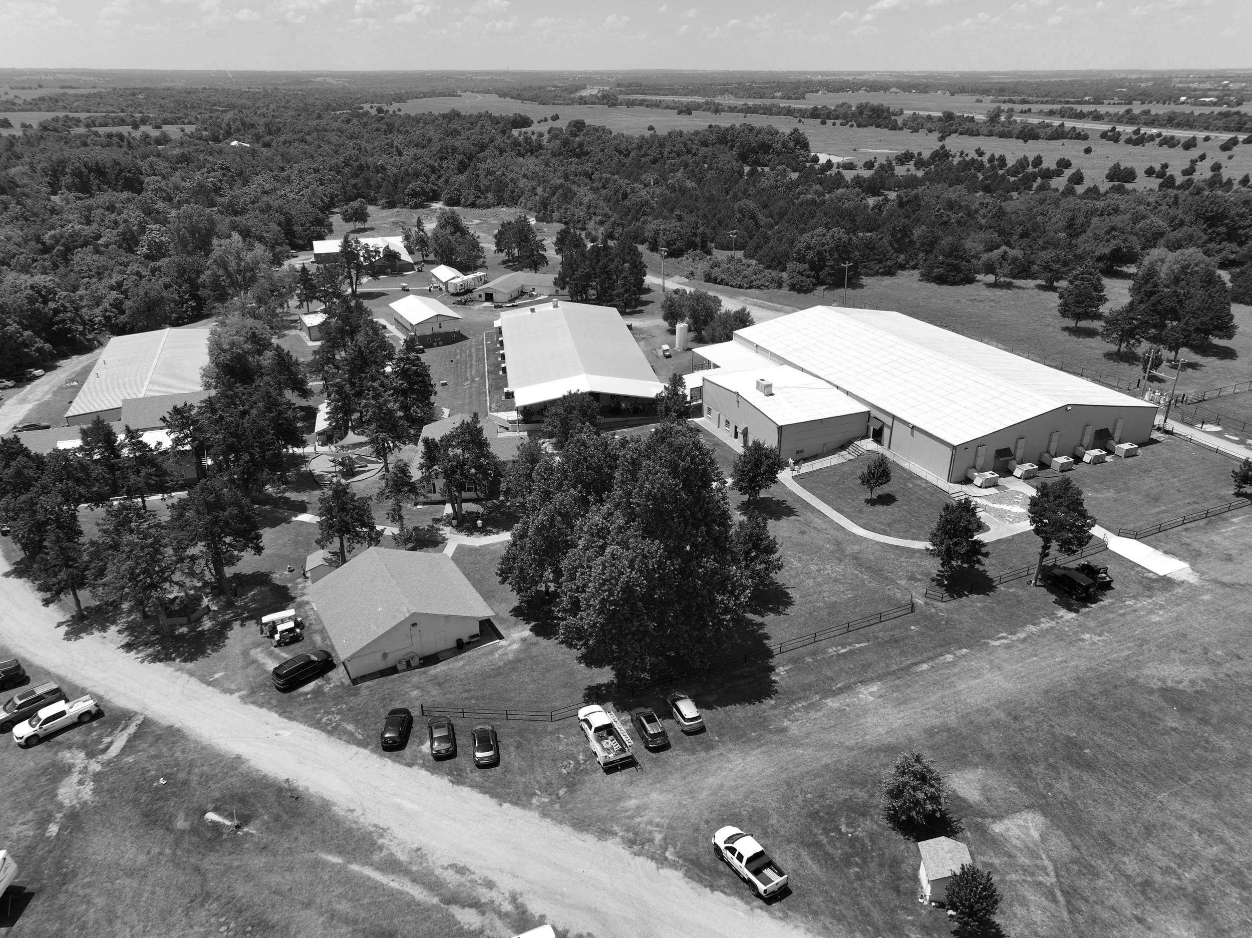 Aerial view of a rural farm with multiple buildings, trees, and a dirt parking area surrounded by open fields and woods.