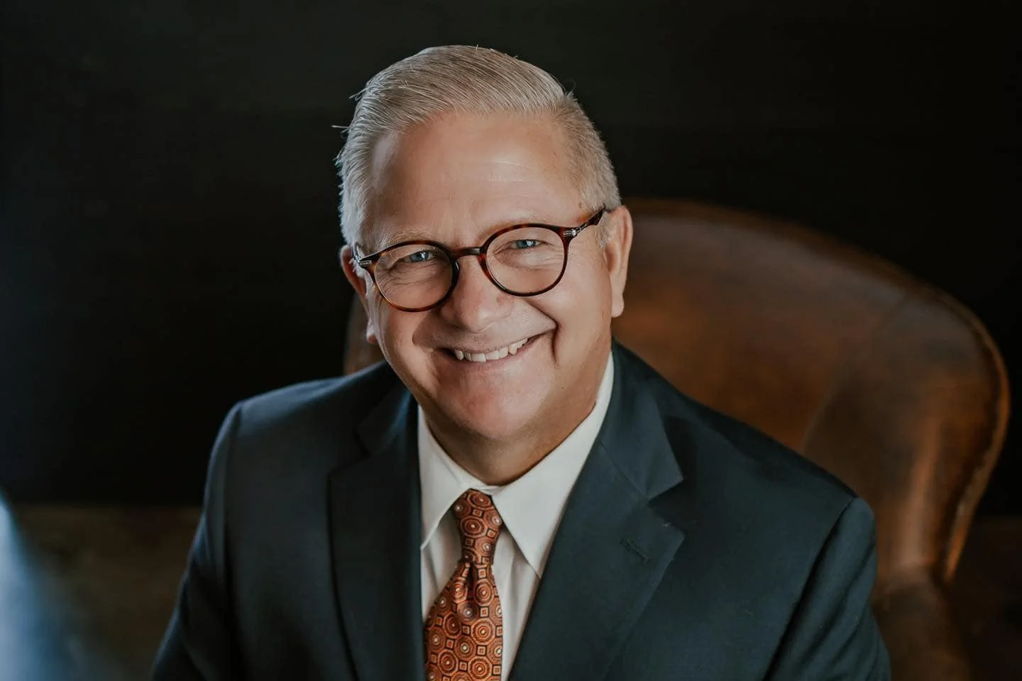 A smiling middle-aged man with glasses, wearing a suit and tie, sitting in a leather chair.