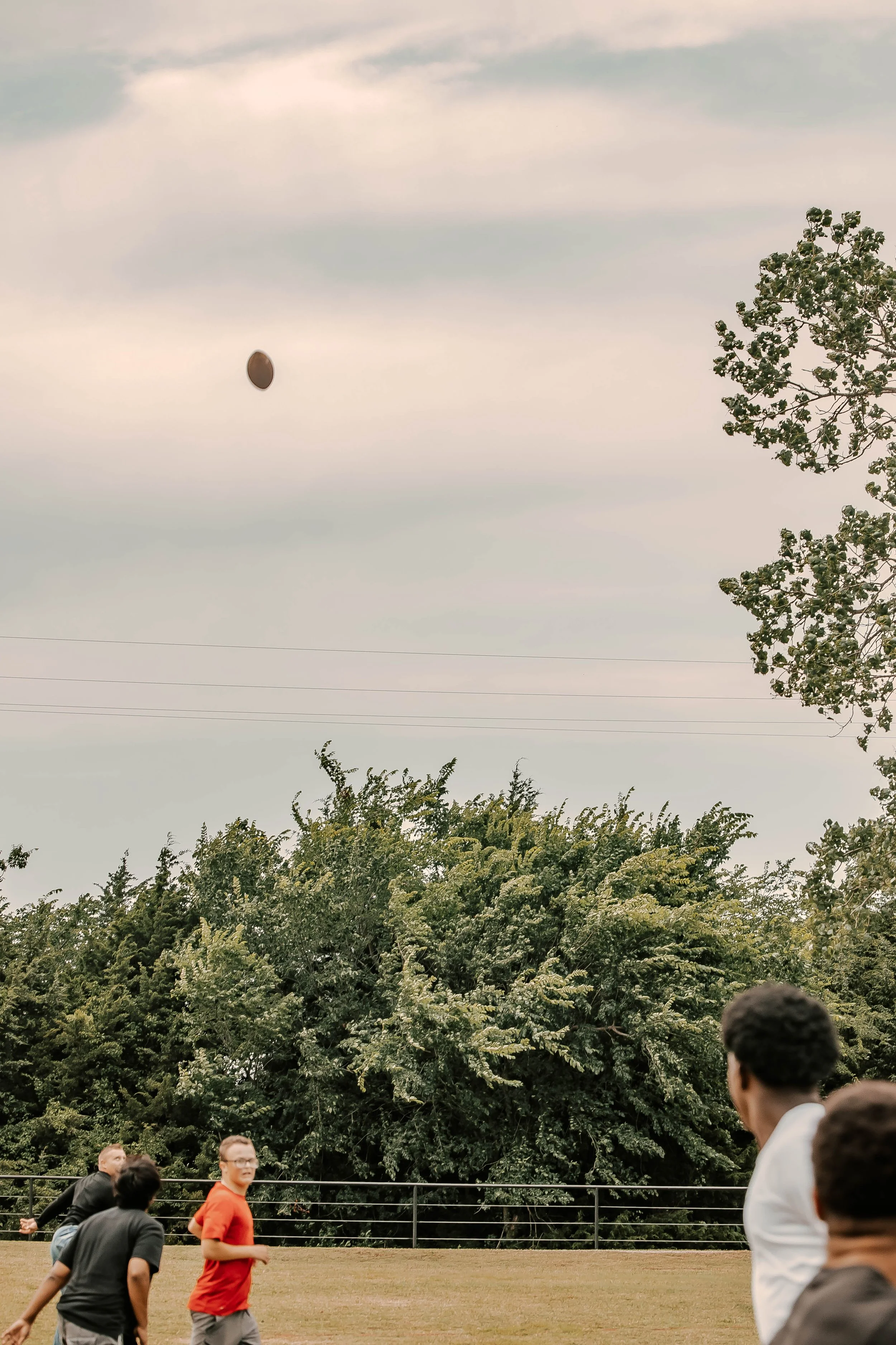 People playing football outdoors on a grassy field with trees and a fence in the background, as a football is in mid-air.