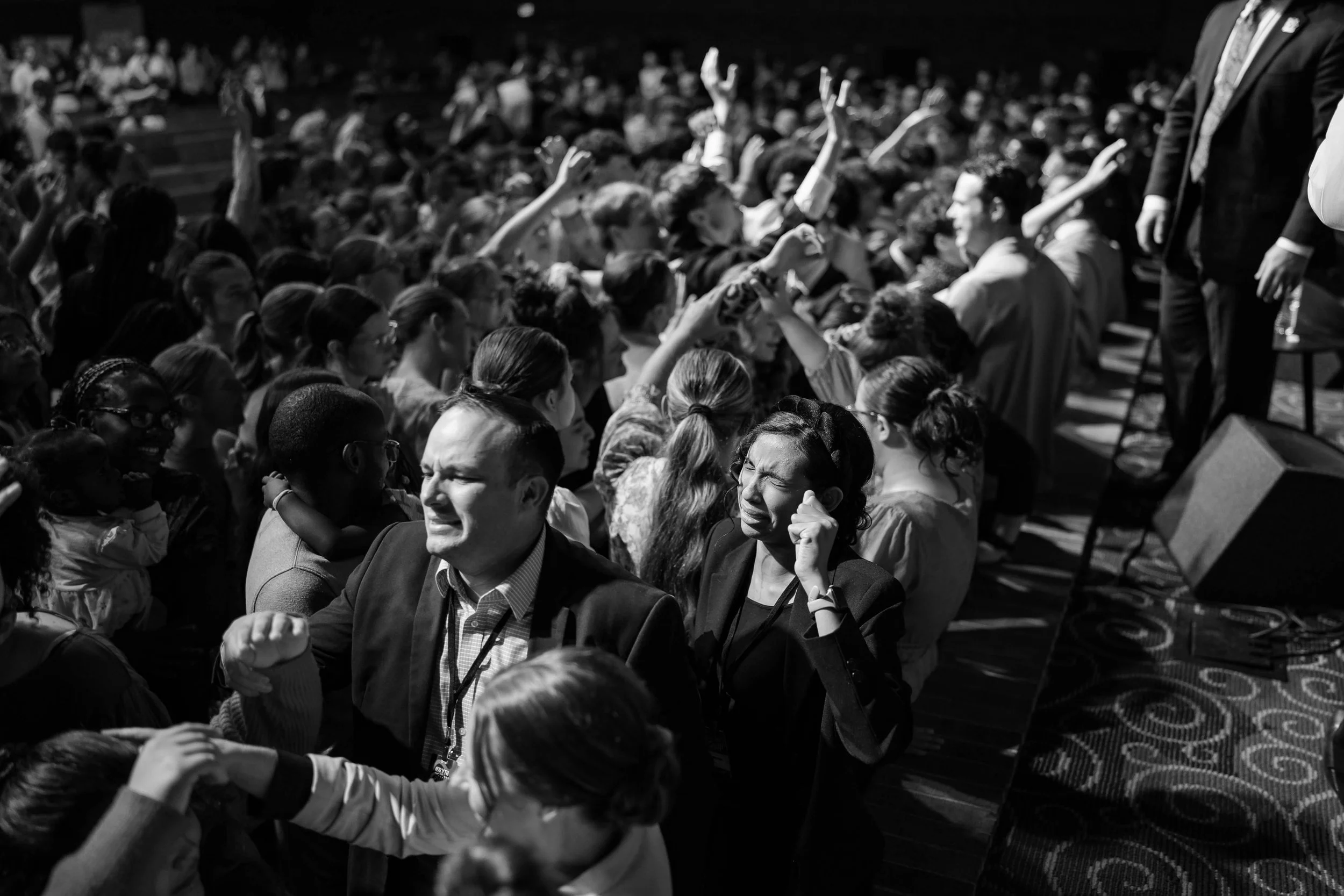 A black and white photo capturing a large gathering of people, some raising their hands, in a conference or seminar setting. The audience appears engaged, with a mix of adults and children.
