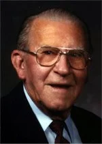 Portrait of an elderly man wearing glasses, a dark blazer, and a light shirt.