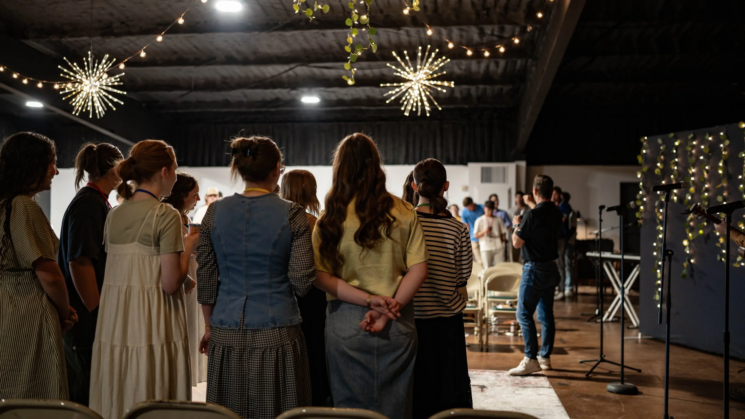 A group of people standing together in a dimly lit room, possibly attending a gathering or event, with decorative lighting and a stage area in the background.