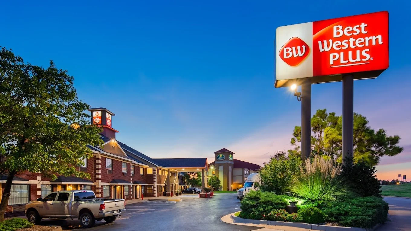 Exterior of a BW Best Western Plus hotel at dusk with a large illuminated sign, brick hotel buildings, parked vehicles, trees, and landscaped greenery.