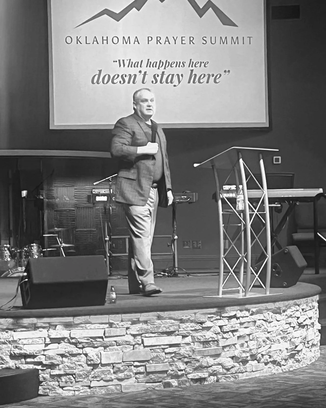 A man holding a microphone standing on a stage with a large screen behind him displaying the text 'Oklahoma Prayer Summit' and the quote 'What happens here doesn't stay here'. The stage has a stone brick front and various musical equipment.